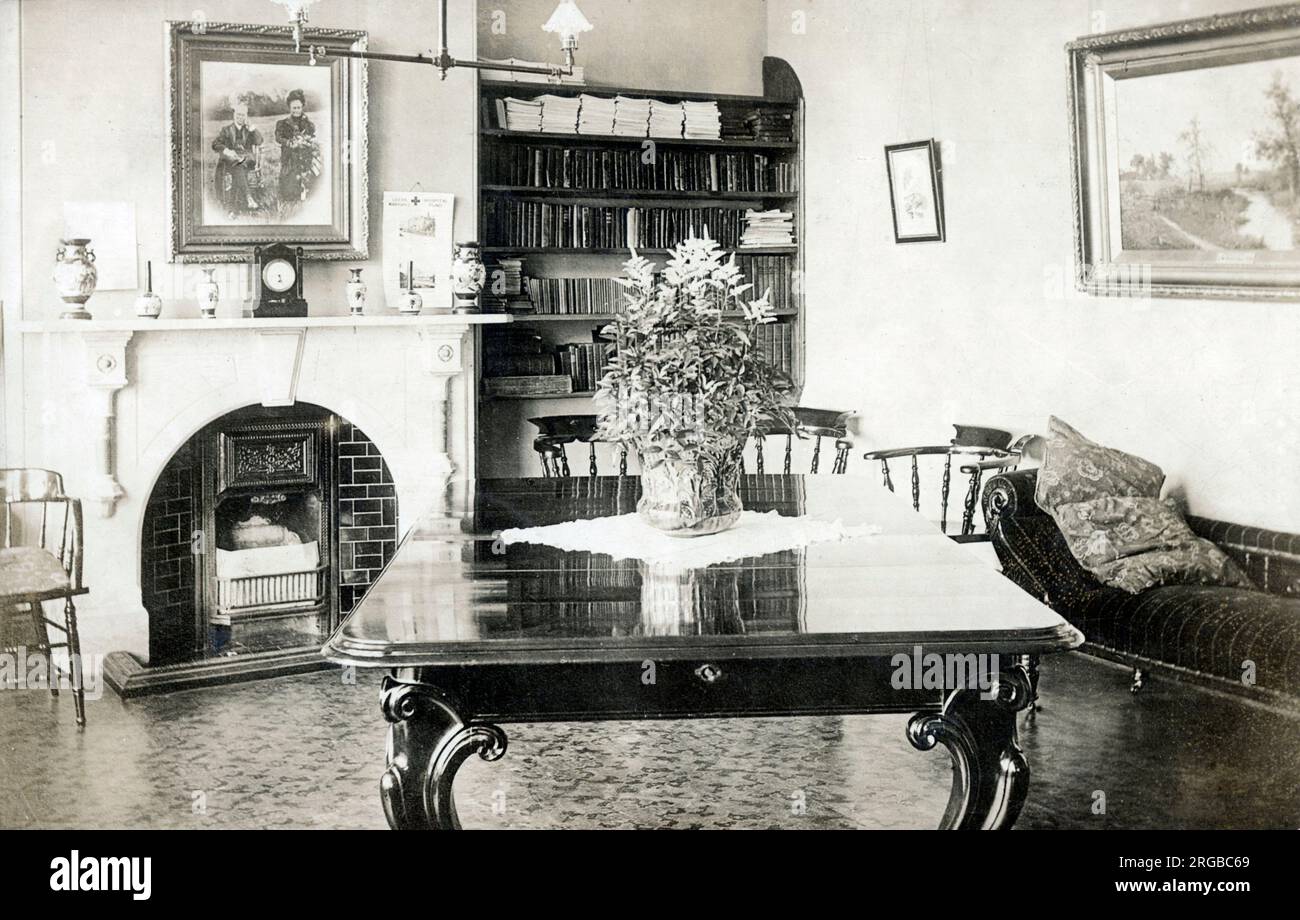 Élégante salle de réception édouardienne avec table en bois poli, chaise longue, grand portrait photographique de famille au-dessus de la cheminée et étagères soignées aux volumes en cuir. Banque D'Images