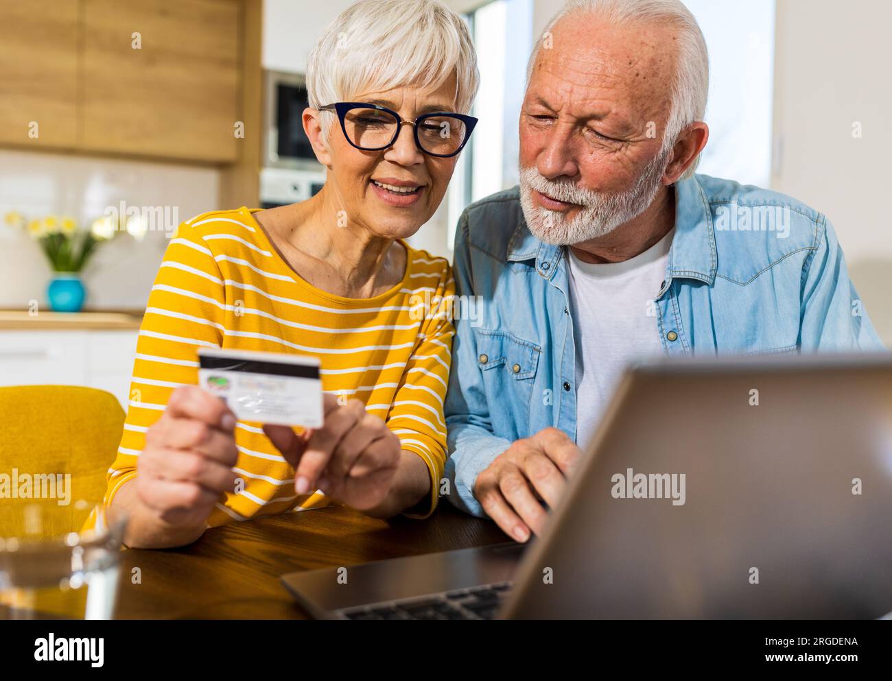 Joyeux couple senior assis à la table de salle à manger avec cuisine en arrière-plan et acheter en ligne avec carte de crédit Banque D'Images