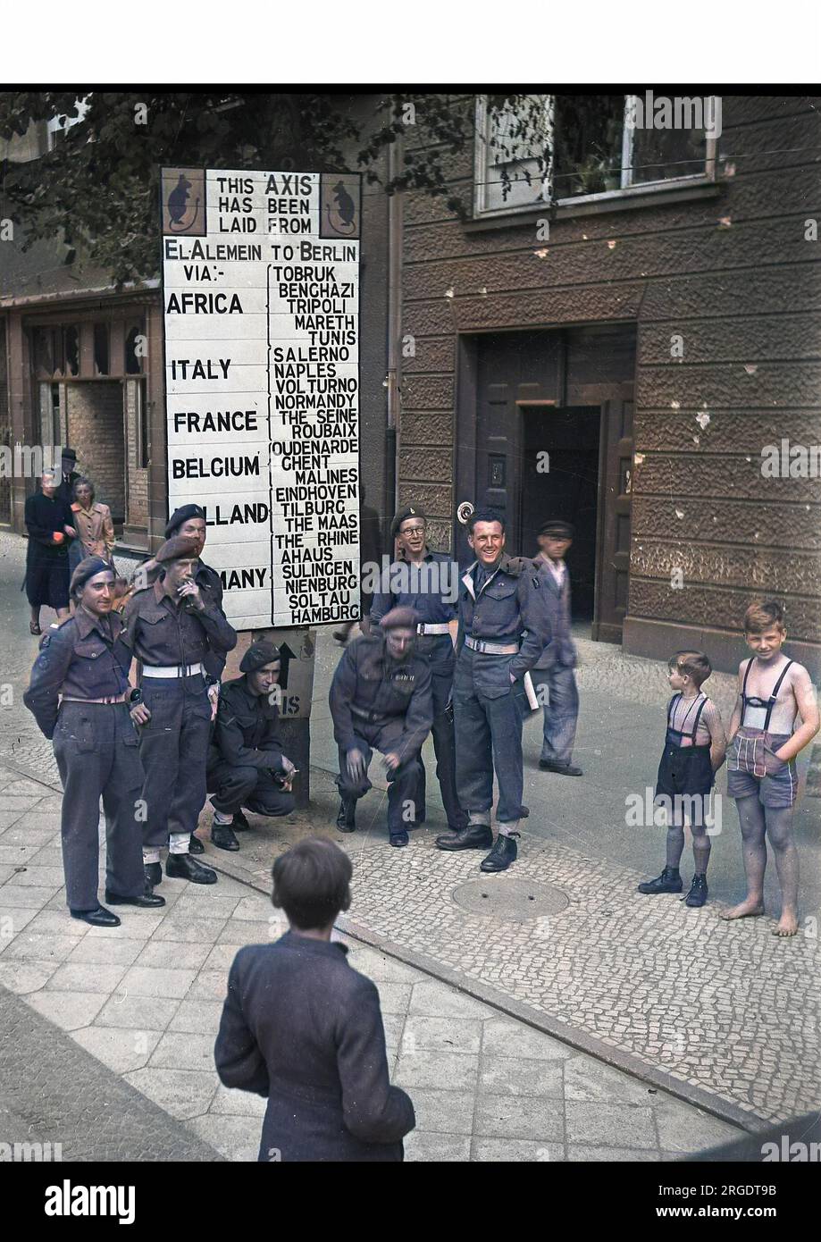 Scène de rue à Berlin, en Allemagne, à la fin de la Seconde Guerre mondiale, avec des soldats et des enfants. Un panneau indique: Cet axe a été posé de El Alamein à Berlin via l'Afrique, l'Italie, la France, la Belgique, la Hollande, Allemagne, avec divers noms de lieux. Banque D'Images