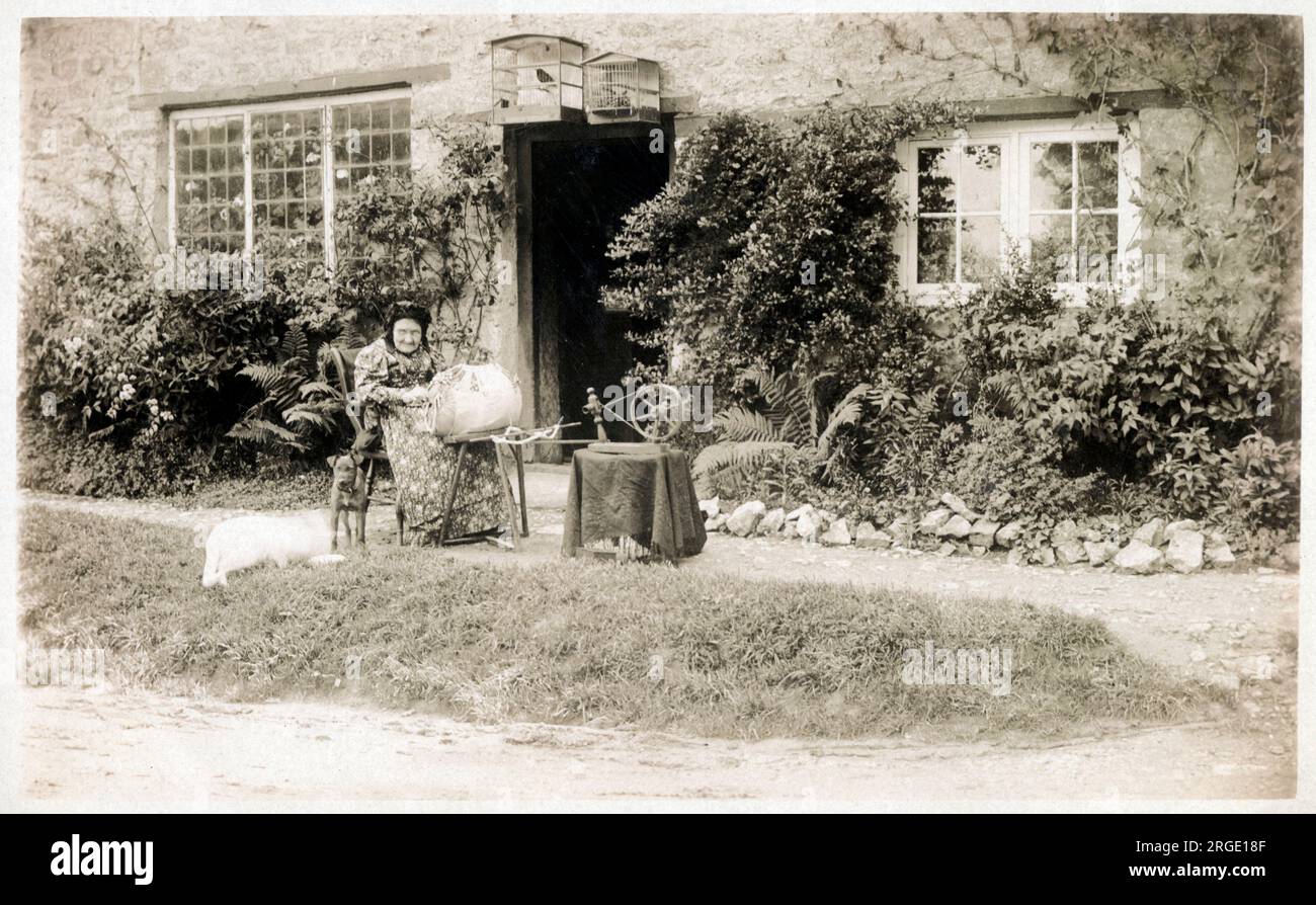 Une dame âgée (entourée de ses nombreux animaux) dentelle faisant à l'extérieur de son charmant chalet rural. Une petite roue qui tourne se tient à côté. Banque D'Images