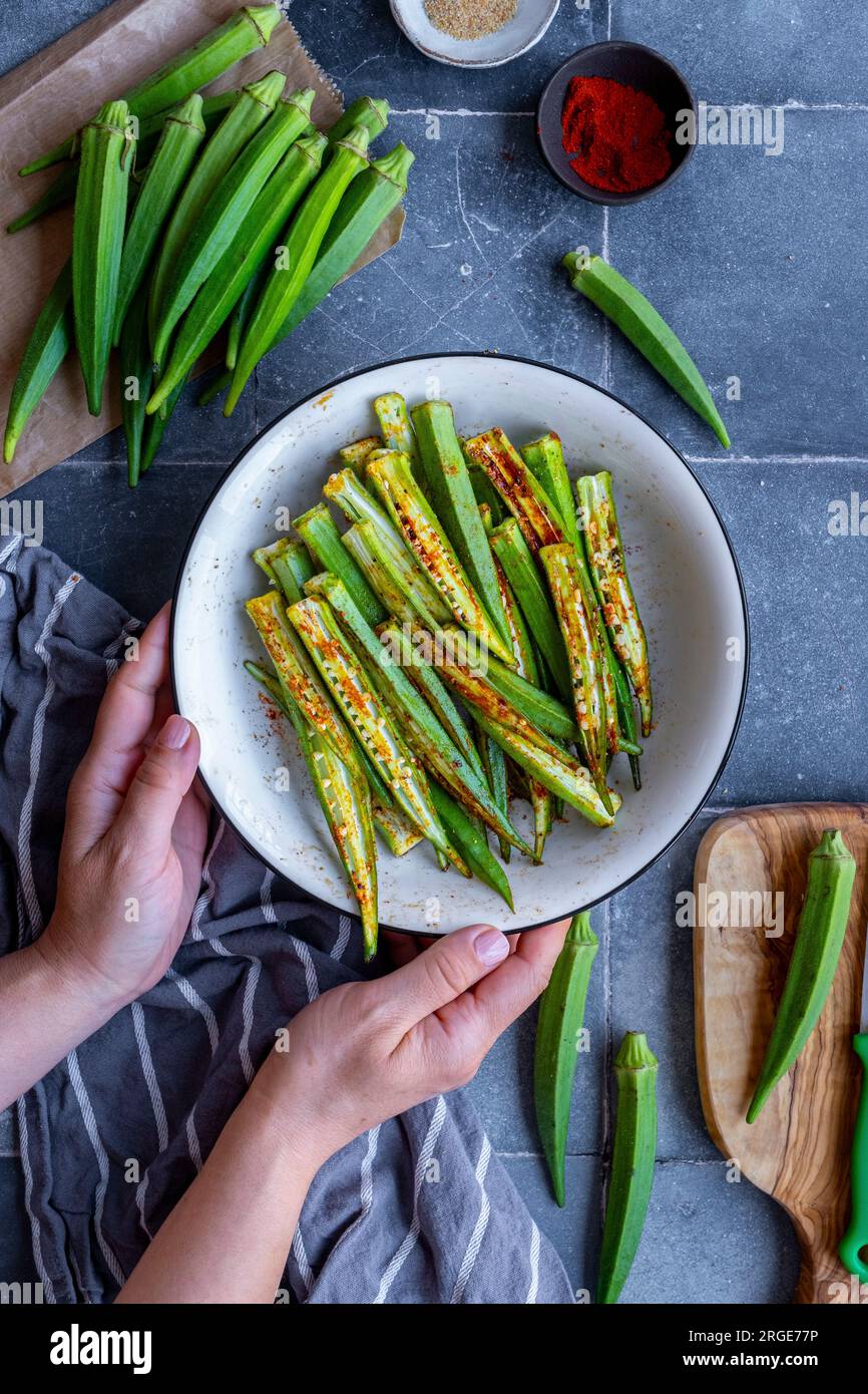 Mains de femme tenant un bol blanc rempli de tranches d'okra sous forme de frites, gousses d'okra entières autour. Banque D'Images