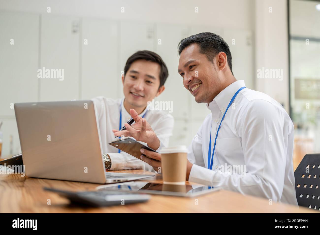 Deux banquiers masculins asiatiques joyeux et professionnels ou analystes financiers regardent un écran d'ordinateur portable et travaillent ensemble au bureau. Banque D'Images