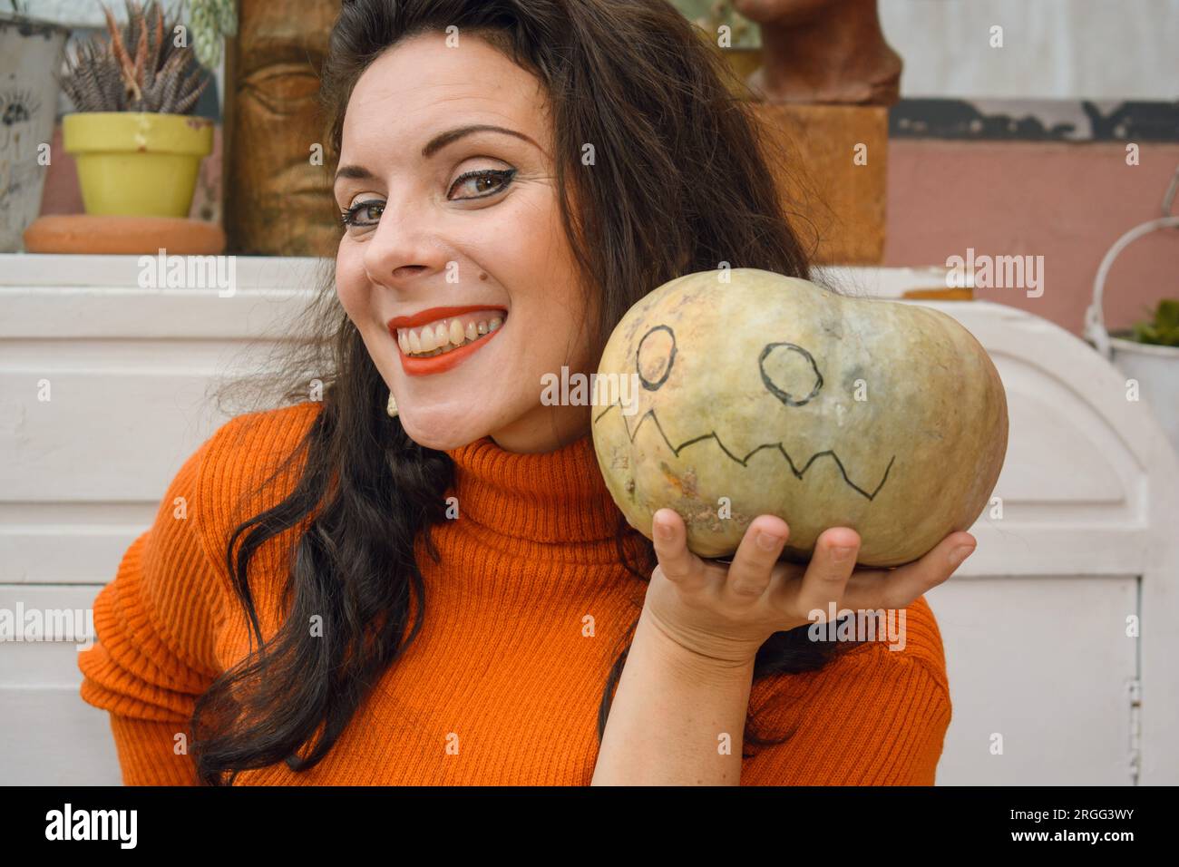 femme caucasienne à la maison en chandail orange, souriante heureuse, debout tenant et montrant la citrouille qu'elle peint avec marquage et décoration pour ha Banque D'Images