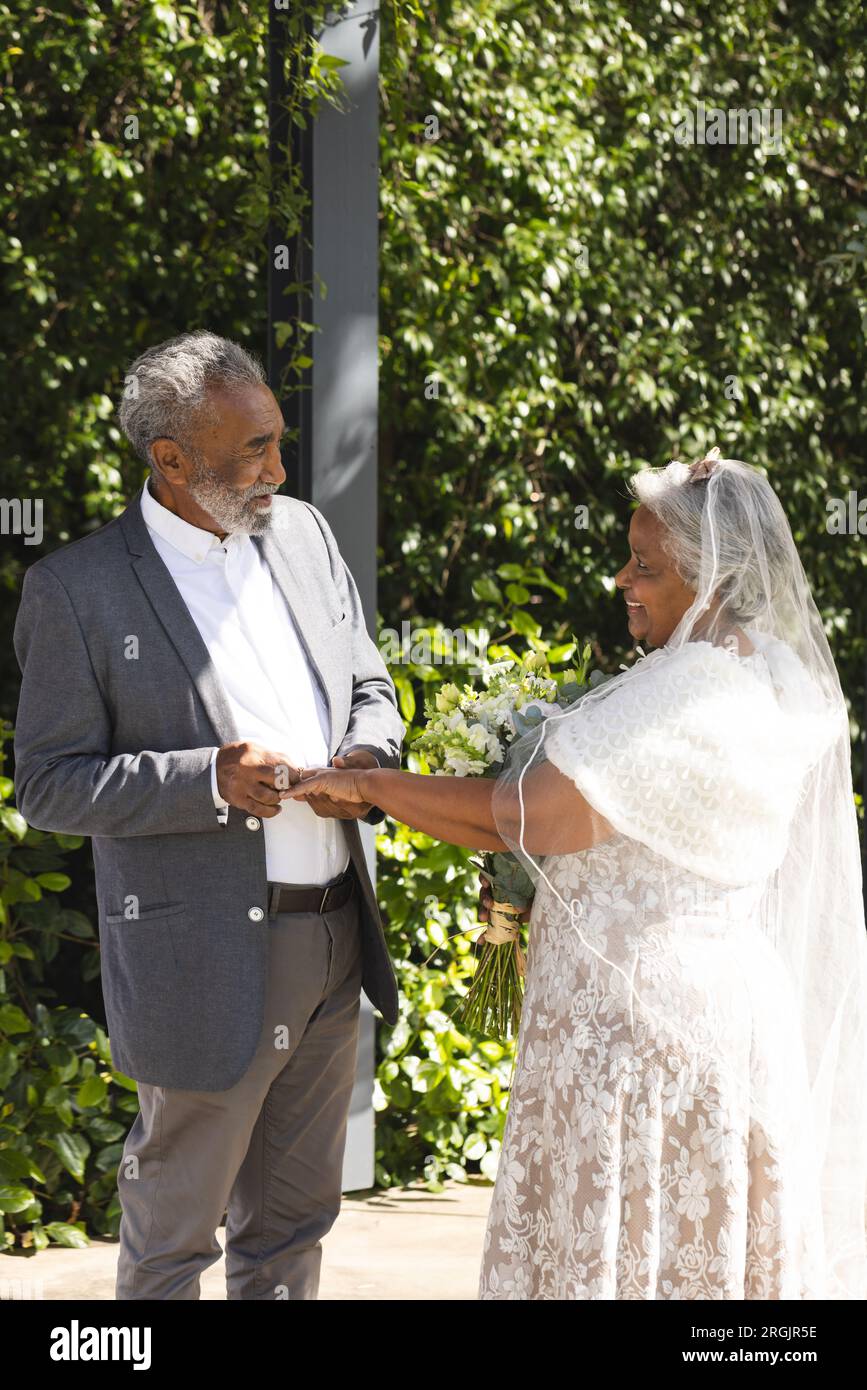 Heureux marié biracial senior mettant la bague sur le doigt de la mariée à la cérémonie de mariage en plein air ensoleillé Banque D'Images
