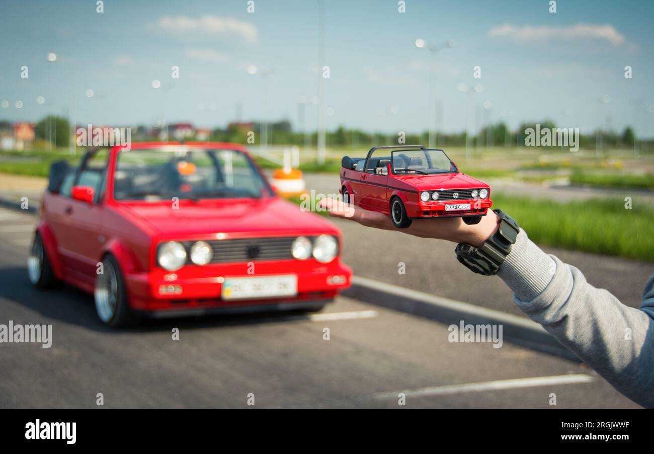 Main à gauche tenant un jouet rouge Volkswagen Golf mk2 cabriolet et une vraie voiture pleine grandeur debout à l'arrêt en arrière-plan. Banque D'Images