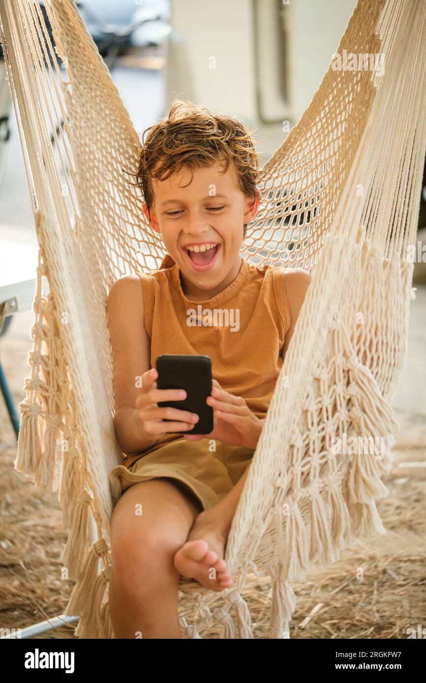 Ravi garçon pieds nus parcourant les médias sociaux sur le téléphone portable et riant à la plaisanterie drôle tout en se relaxant dans le hamac le week-end Banque D'Images