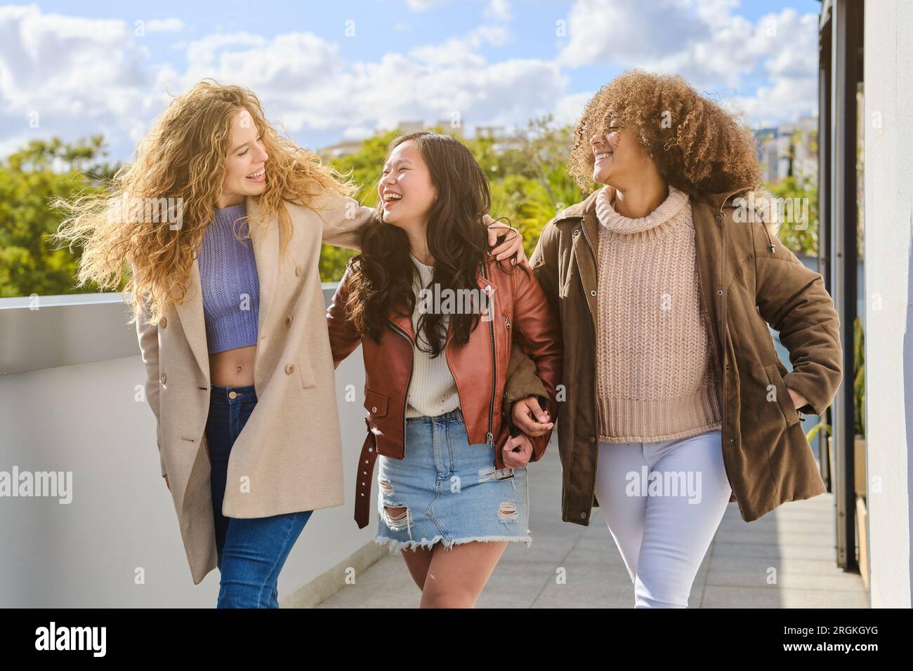 Joyeux jeunes amies diverses riant à la blague et marchant sur la terrasse dans des vêtements chauds en automne tout en se regardant Banque D'Images