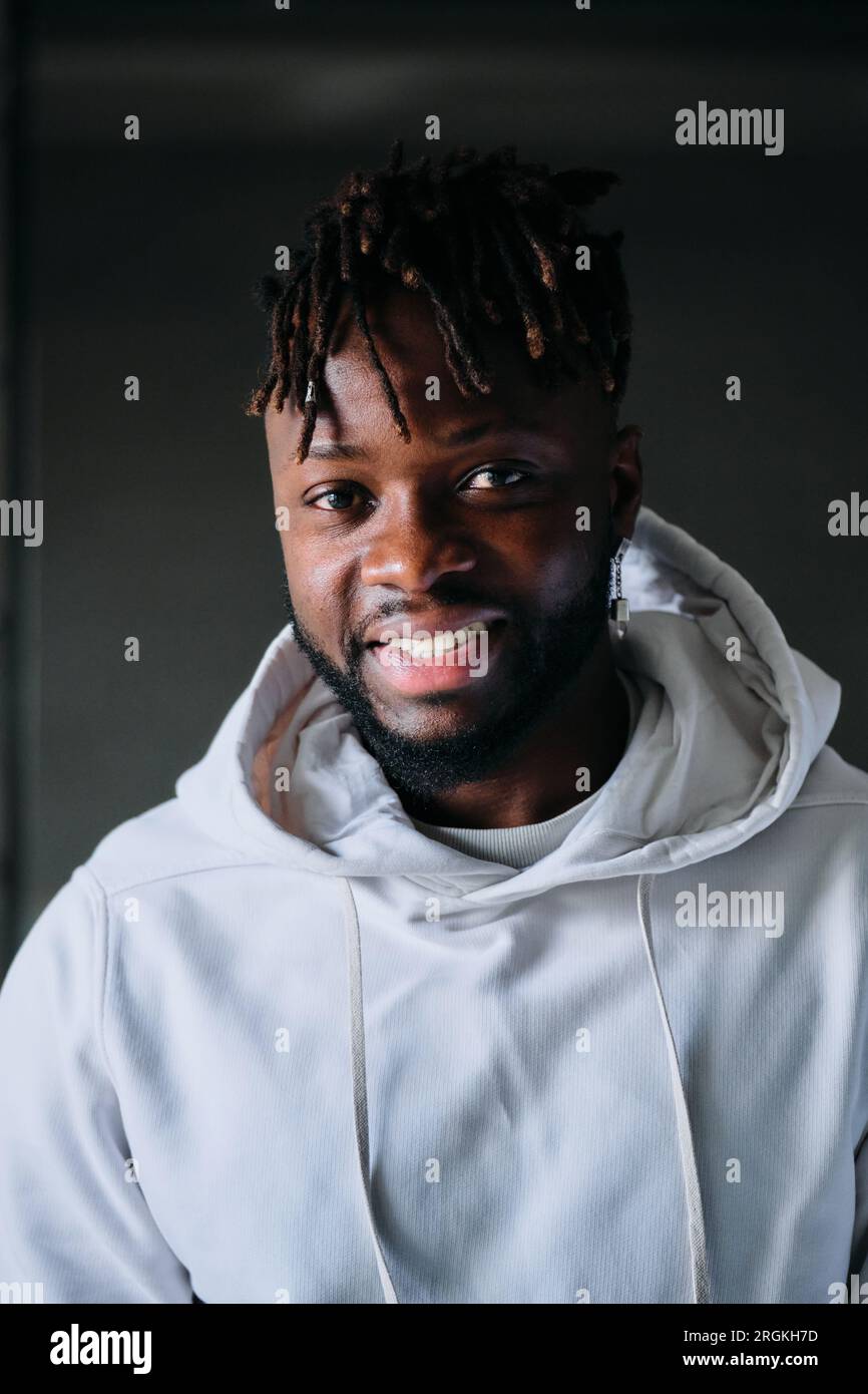 Homme afro-américain élégant confiant dans un sweat à capuche regardant la caméra avec une expression faciale positive tout en se tenant sur un fond sombre dans la pièce Banque D'Images