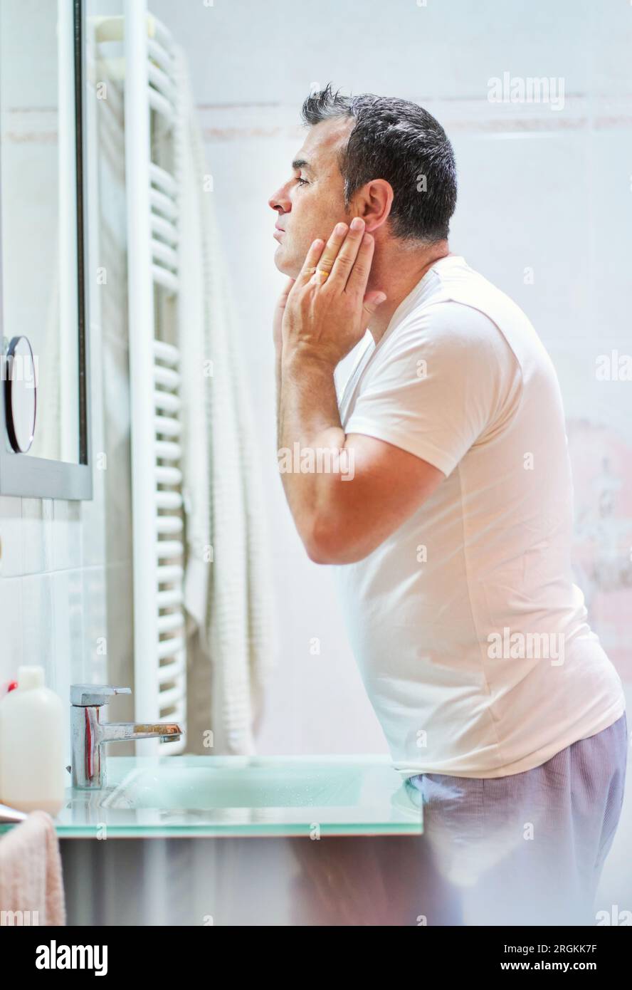 Vue latérale d'un homme mature portant des vêtements domestiques caressant  le visage avec de la crème pendant la routine hygiénique dans la salle de  bain tout en se regardant dans le miroir