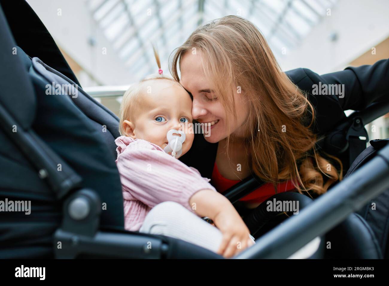 Jeune jolie femme avec un petit enfant dans une voiture. Mère heureuse embrassant son bébé blonde dans des vêtements roses. Maman s'assit à côté de la poussette et discuta Banque D'Images