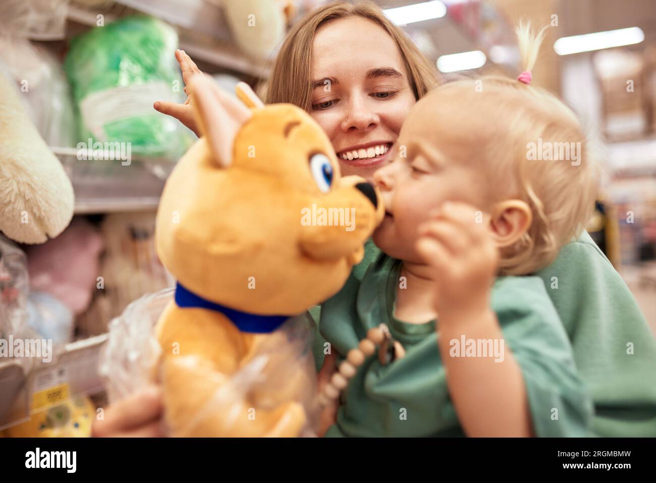 Heureuse jeune mère avec bébé mignon dans ses mains marchant autour du magasin de jouets dans le centre commercial. Maman et petite fille blonde choisissant des jouets et s'amusant. Banque D'Images