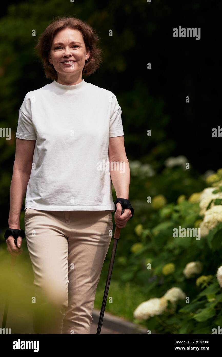 Femme mature attrayante dans le parc avec bâtons de marche nordique. Femme brune d'âge moyen en t-shirt blanc marchant dans la ruelle du parc. Garçon senior Banque D'Images