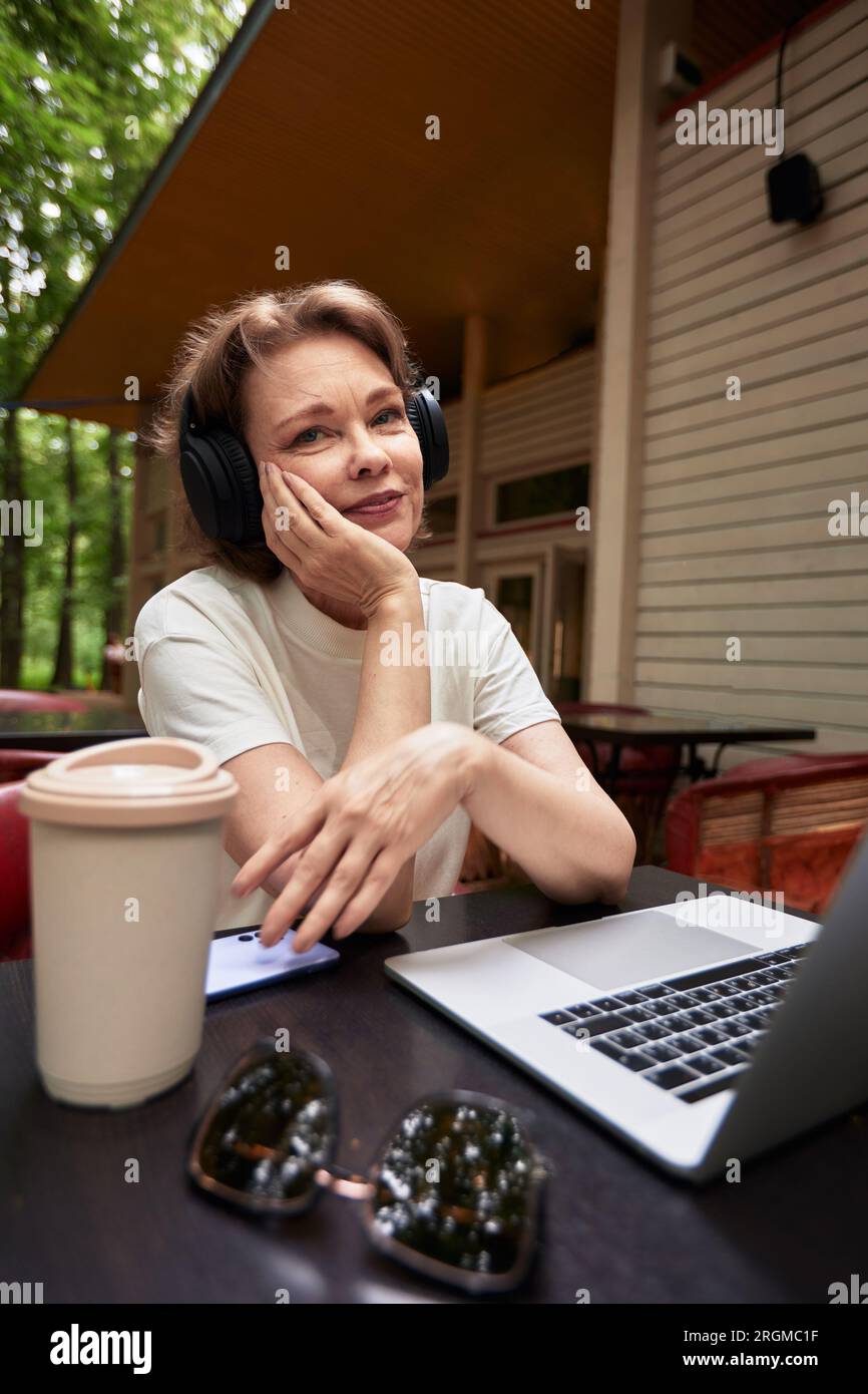 Adorable femme mature avec ordinateur portable et écouteurs sans fil assis à la table dans le café en plein air. Brunette souriante d'âge moyen en t-shirt blanc utilisant Mo Banque D'Images