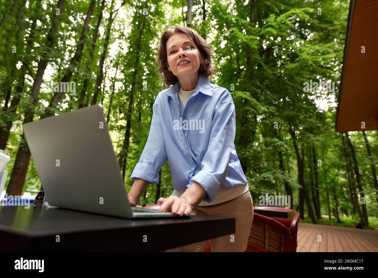 Adorable femme mature avec ordinateur portable assis à la table dans le café en plein air dans le parc. Brunette souriante d'âge moyen dans une chemise bleue fonctionne et joue en utilisant Banque D'Images
