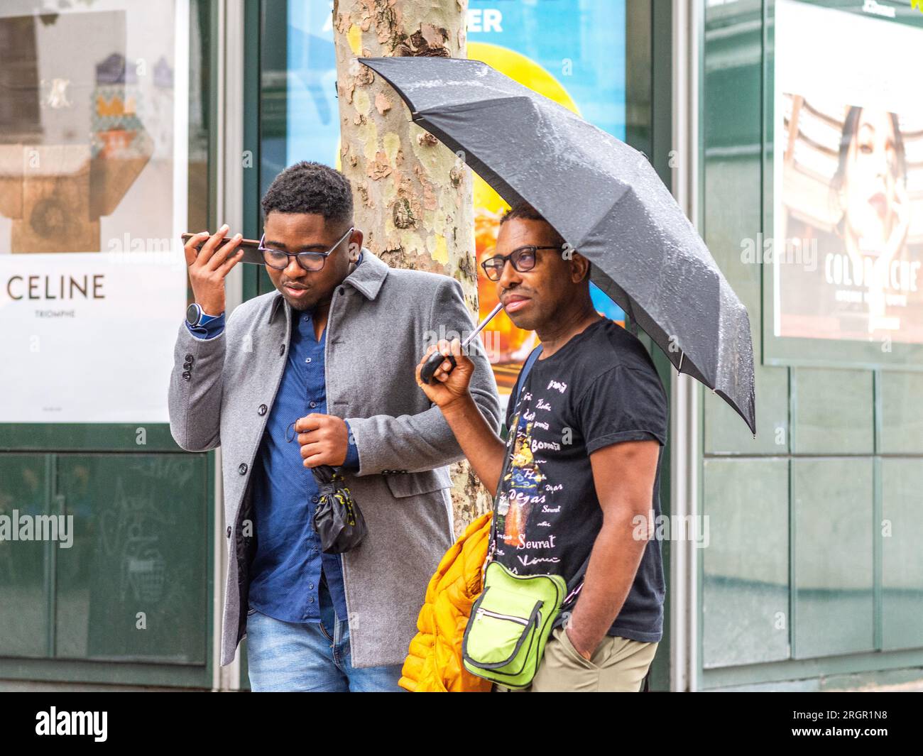 Deux amis masculins noirs, l'un tenant un parapluie l'autre faisant un appel téléphonique dans Paris pluvieux, France. Banque D'Images