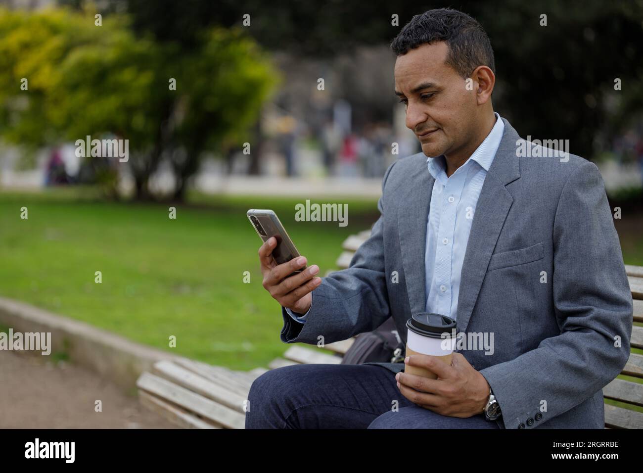 Portrait d'homme latino en veste regardant un téléphone portable assis sur un banc dans un parc public avec espace de copie. Banque D'Images