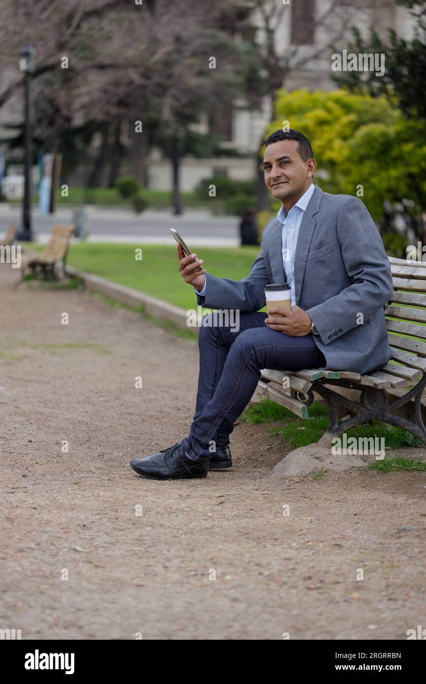 Homme latino en veste avec une tasse de café jetable assis sur un banc dans un parc public. Banque D'Images