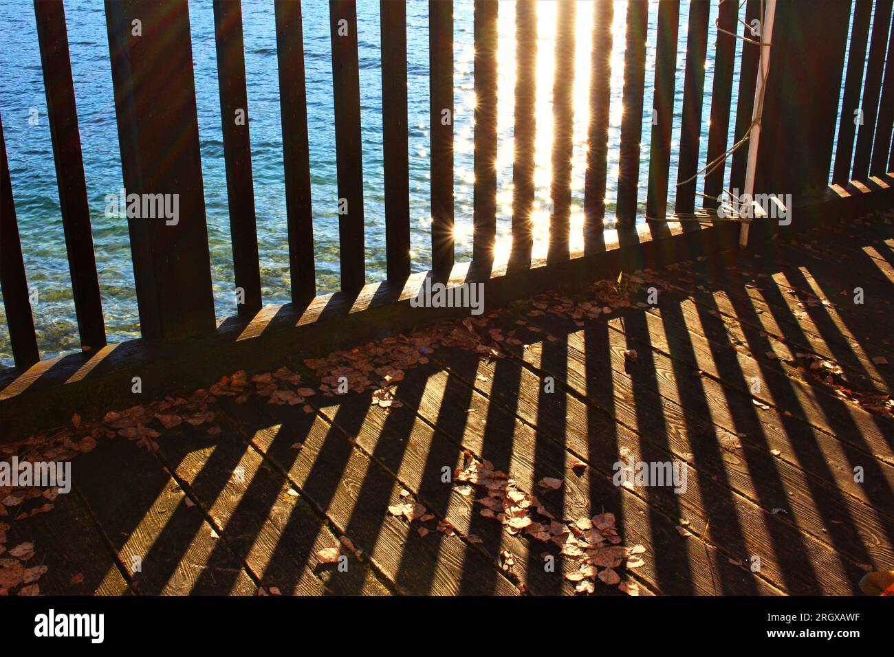 Soleil se reflétant sur le lac et brillant sur une clôture en bois projetant des ombres sur la terrasse en bois. Fond de soirée d'été frais et symétrique Banque D'Images