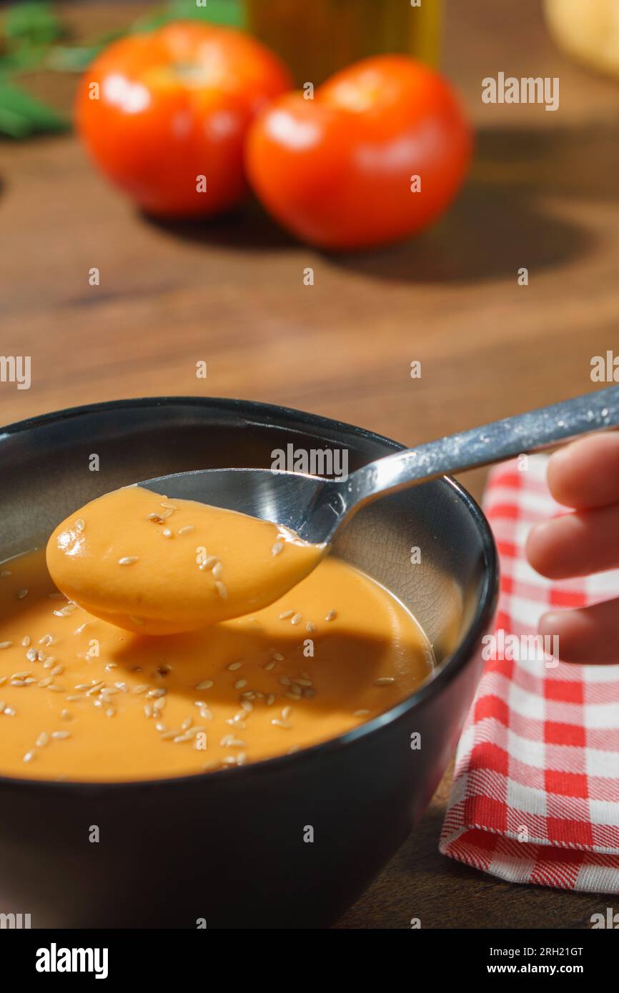 Bol de gazpacho andalou espagnol, aux graines de sésame, sur une table en bois sombre avec une serviette à carreaux rouges, une cuillère, des tomatos et de l'huile d'olive. Banque D'Images