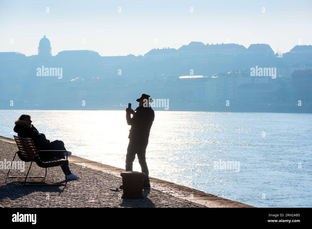 Silhouette d'homme prenant la photo d'une femme assise sur le banc à la rivière contre la ligne d'horizon de la ville Banque D'Images