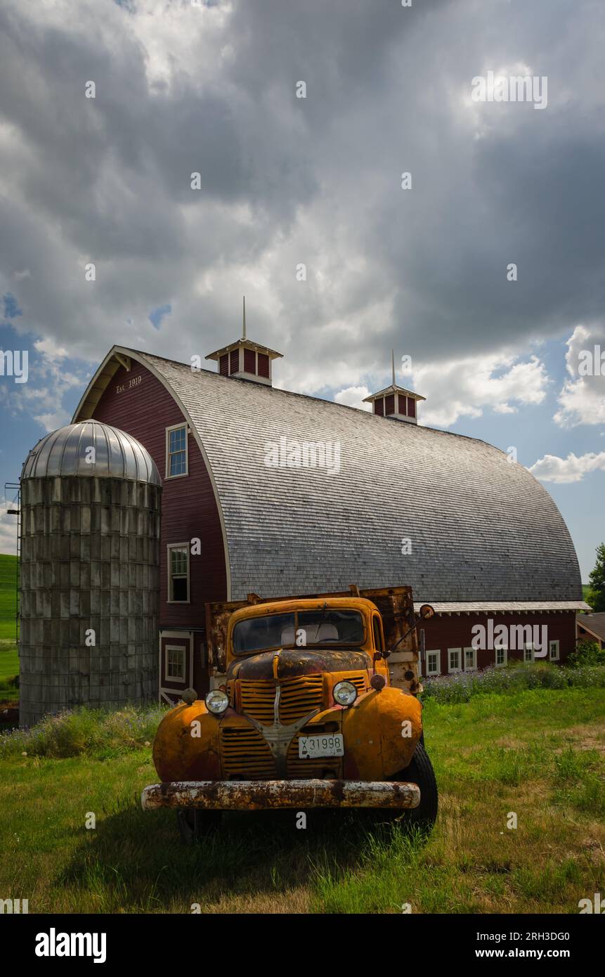 Un vieux camion orange rouillé et une grande grange rouge (construite en 1919) avec toit en cèdre, deux coupoles et un petit silo. Colfax, Washington, États-Unis. Banque D'Images