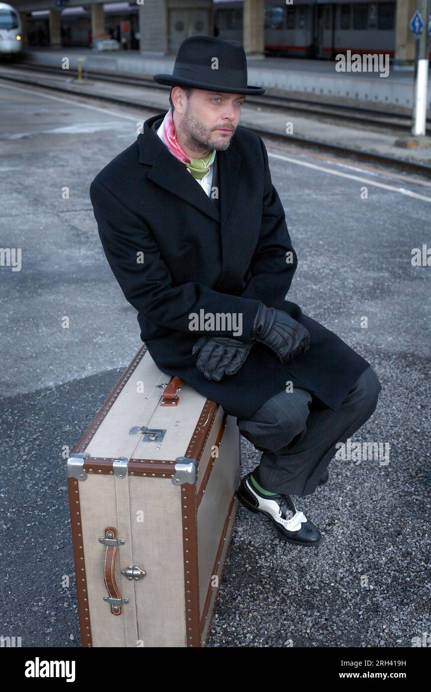 Voyageur masculin attendant de voyager sur le quai de chemin de fer. Homme assis sur des valises à la gare centrale, train à l'heure ou retardé. Banque D'Images