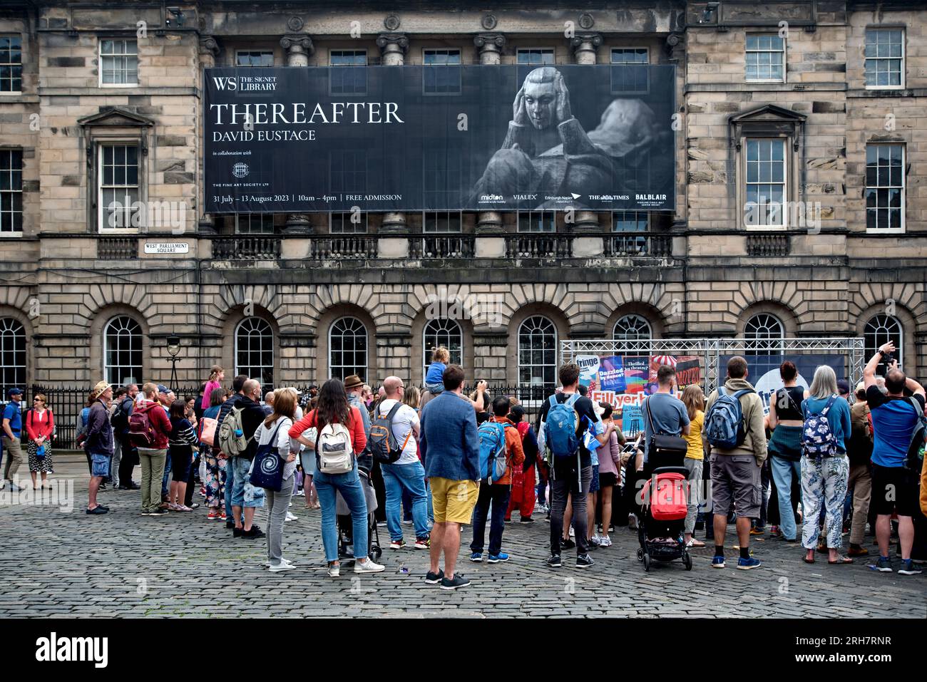 Vue arrière du public regardant un artiste de rue avec une bannière annonçant une exposition David Eustace à la signet Library, Édimbourg, Écosse, Royaume-Uni. Banque D'Images