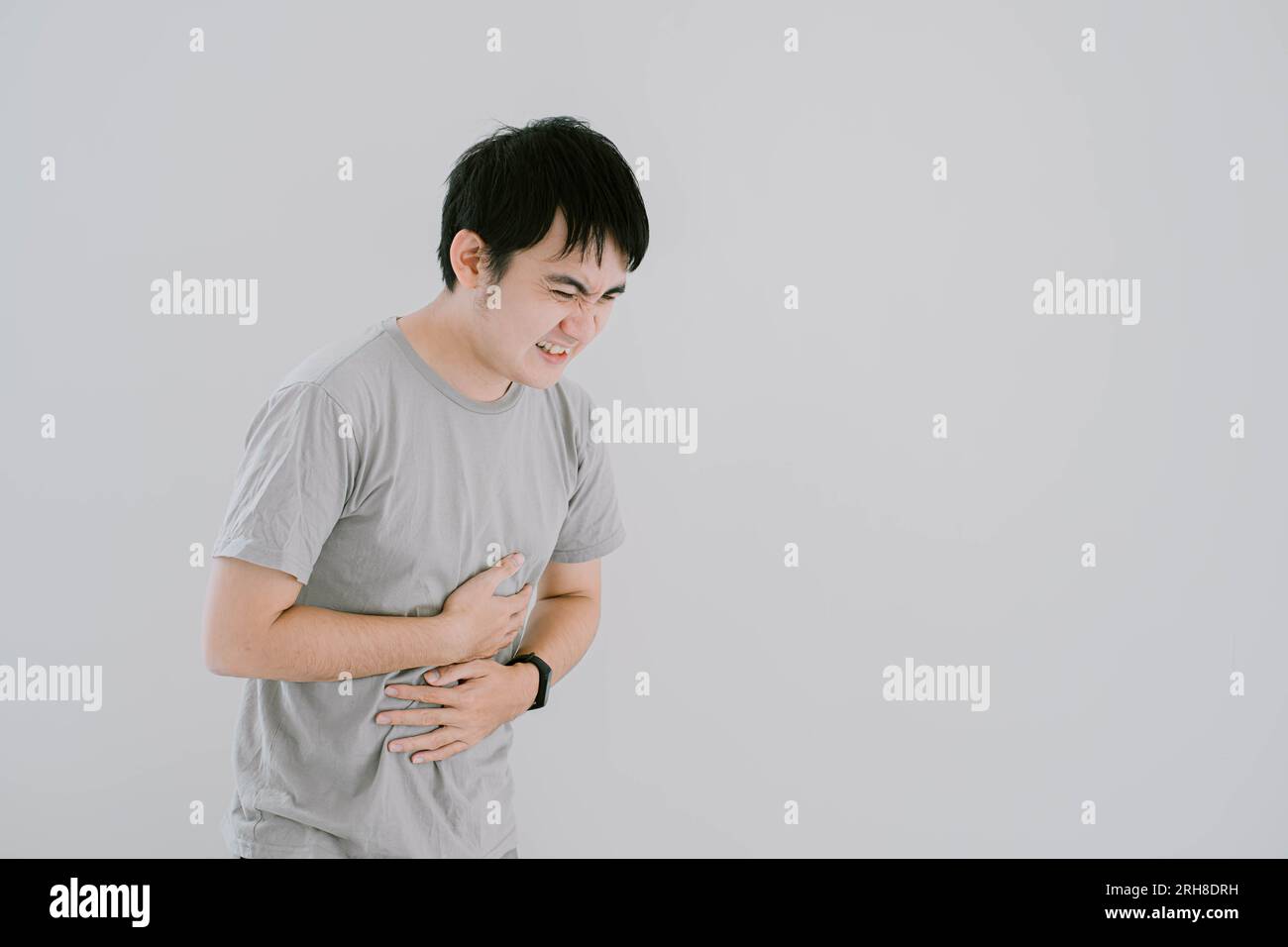 Photo de mise au point sélective d'un jeune homme asiatique portant un T-shirt décontracté vert sauge et une smartwatch ont eu mal au ventre et le tient avec les mains sur une balle blanche isolée Banque D'Images