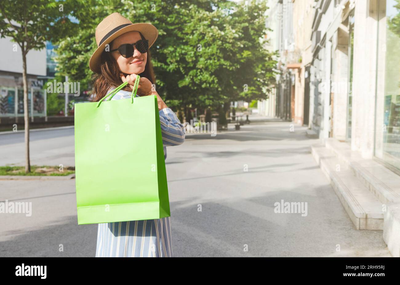 Jeune femme portant un chapeau et des lunettes de soleil magasinant tout en marchant dans la rue, tenant un sac à provisions de couleur verte à la main et souriant. Banque D'Images