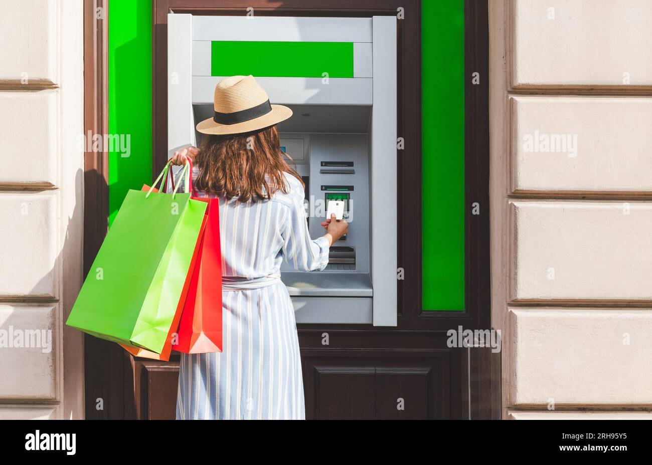 Vue arrière de la femme à la mode dans le chapeau de paille, tenant des sacs à provisions avec ses achats et en utilisant le guichet automatique bancaire dans la rue. Banque D'Images