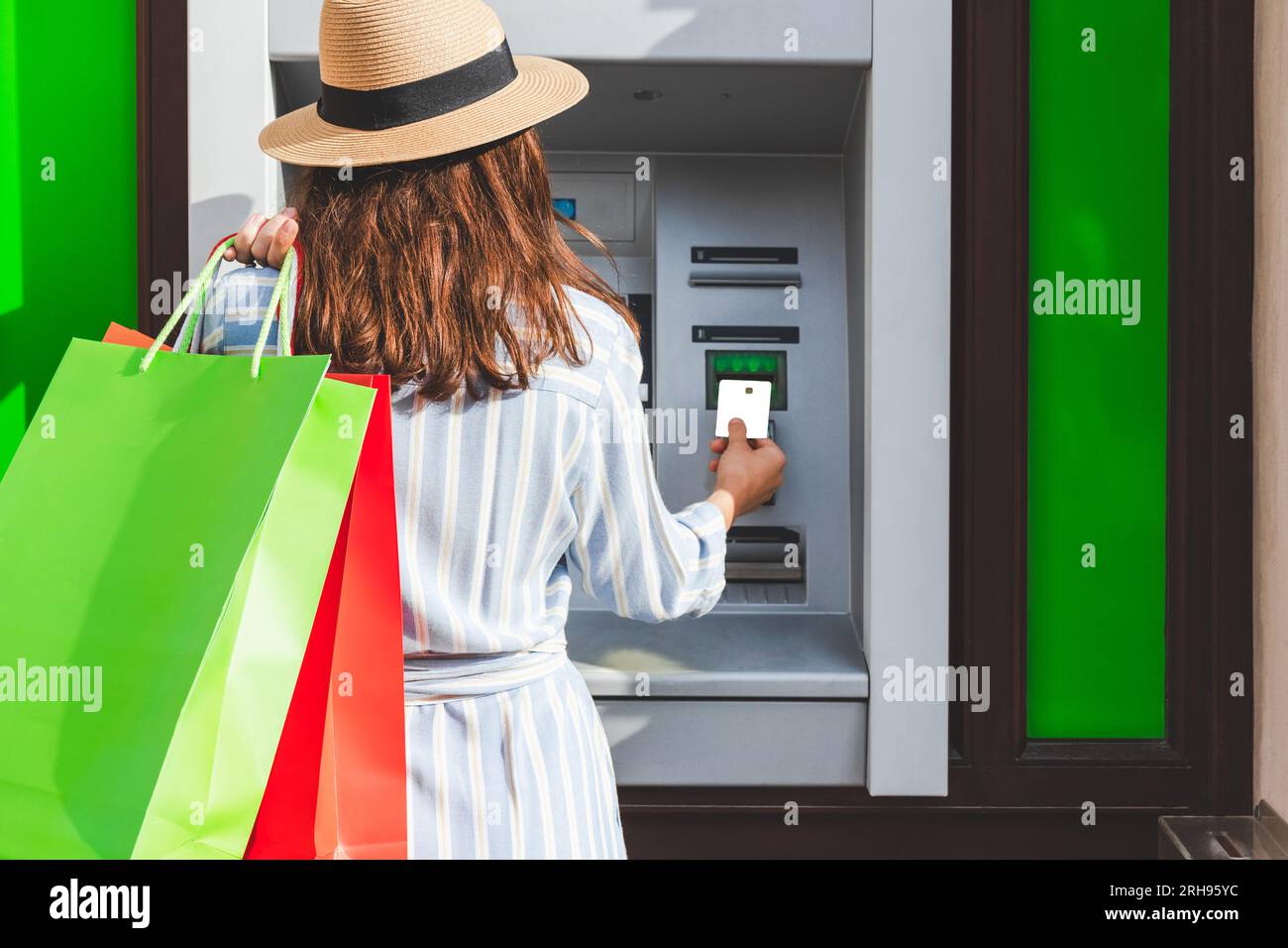 Vue arrière de la femme à la mode dans le chapeau de paille, tenant des sacs à provisions avec ses achats et en utilisant le guichet automatique bancaire dans la rue. Banque D'Images