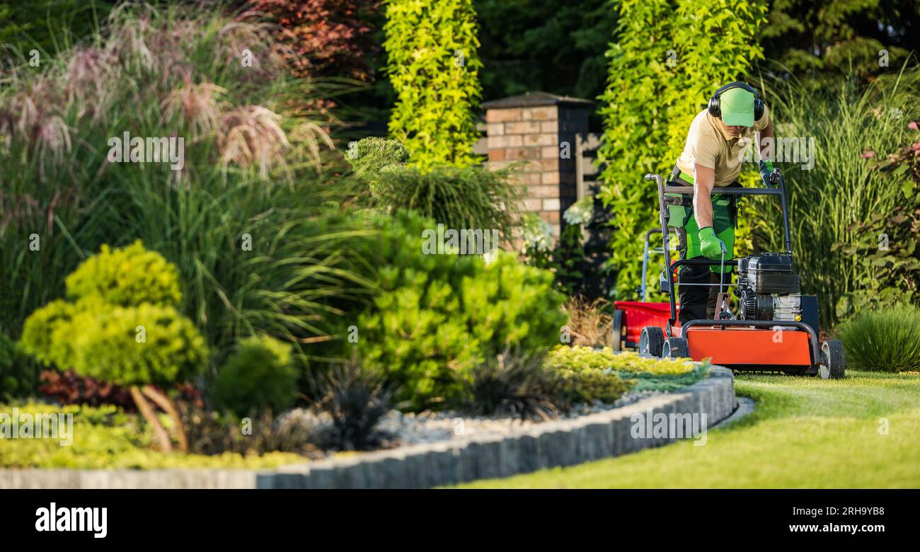 Ouvrier d'aménagement paysager caucasien dans ses années 40 et aération de pelouse à l'aide d'un puissant aérateur alimenté par essence Banque D'Images