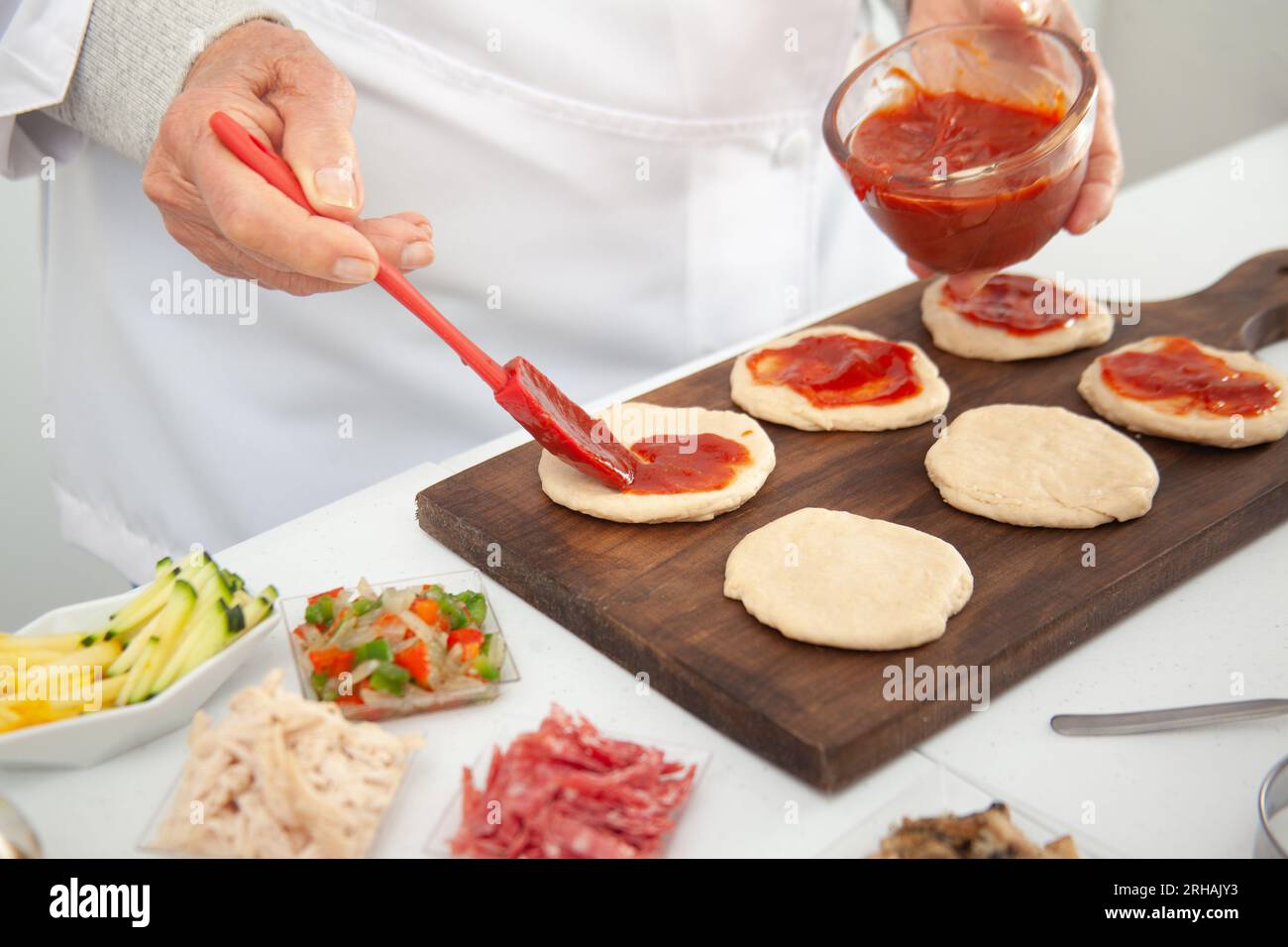 Femme senior ajoutant de la sauce pizza à la pâte étirée. Délicieuse préparation de mini pizzas maison. Banque D'Images