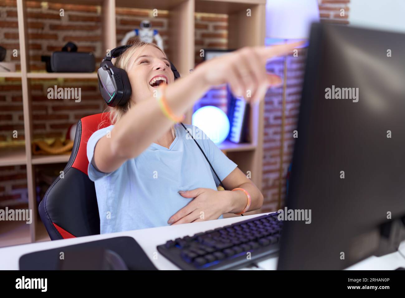 Jeune femme caucasienne jouant à des jeux vidéo portant des écouteurs riant de vous, pointant du doigt la caméra avec la main sur le corps, expression de honte Banque D'Images