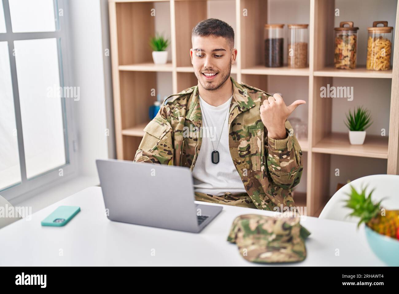 Jeune homme hispanique portant un uniforme militaire de camouflage faisant un appel vidéo pointant le pouce vers le côté souriant heureux avec la bouche ouverte Banque D'Images