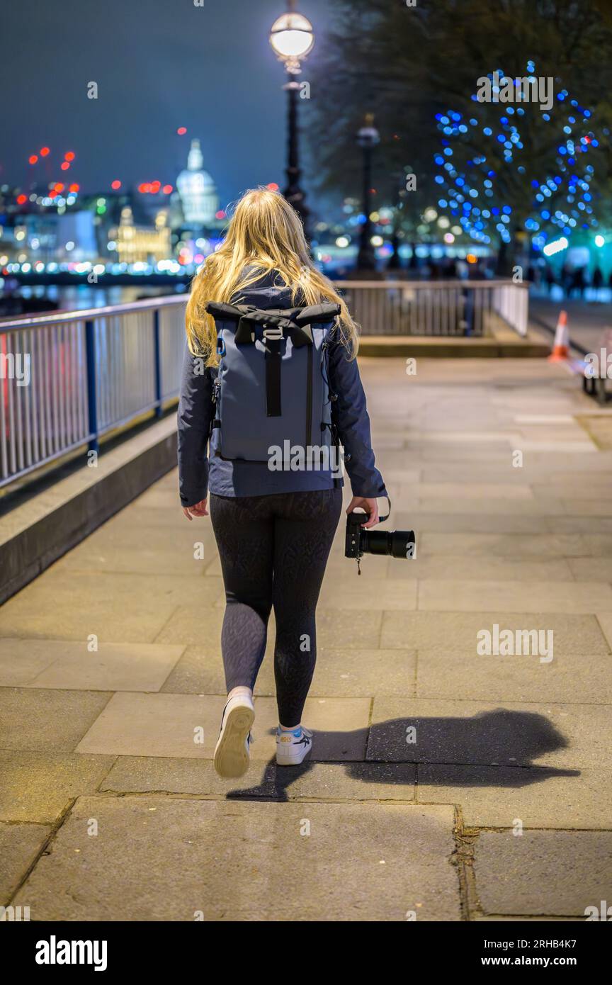 Une photographe blonde marche le long de Southbank la nuit, caméra à la main, avec la cathédrale Saint-Paul brillamment illuminée en arrière-plan. Banque D'Images