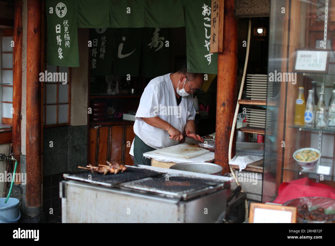 Jour d'été, un vieil homme japonais portant un masque et préparant de la nourriture pour la vente à Fushimi Inari, Kyoto, avec des brochettes à l'avant du magasin Banque D'Images