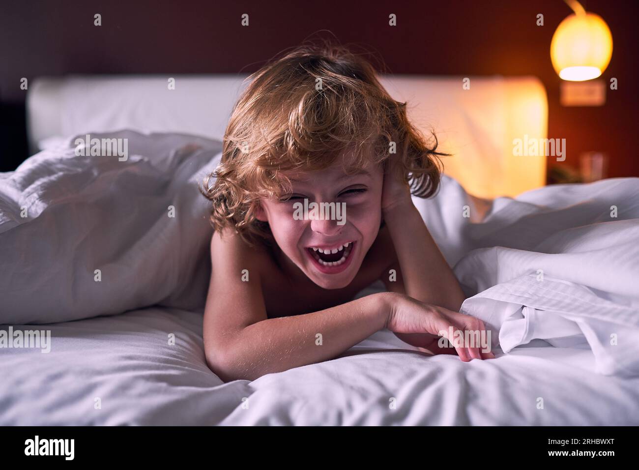 Heureux petit garçon touchant les cheveux et riant à la blague tout en se reposant sous la couverture sur un lit moelleux dans l'hôtel Banque D'Images