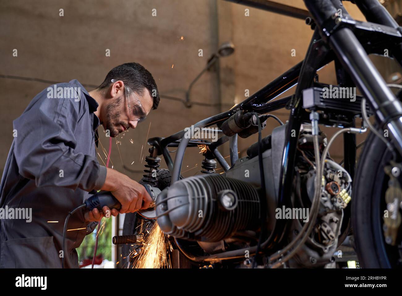 Vue latérale du technicien barbu en uniforme et lunettes de protection fixant la moto avec meuleuse d'angle en atelier Banque D'Images