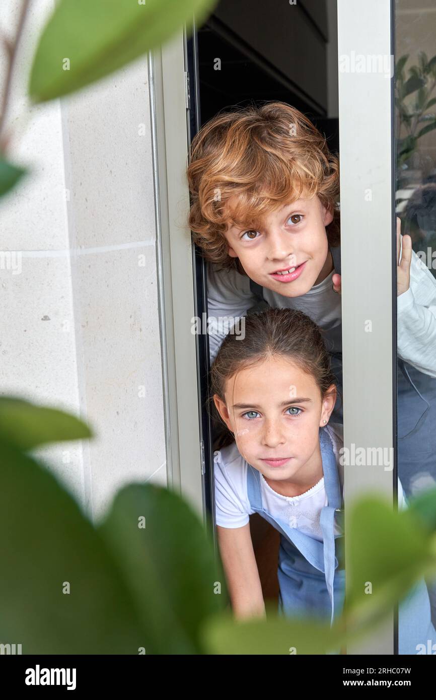 Garçon avec les cheveux bouclés et fille en jeans dans l'ensemble regardant hors de la porte en verre de la maison Banque D'Images