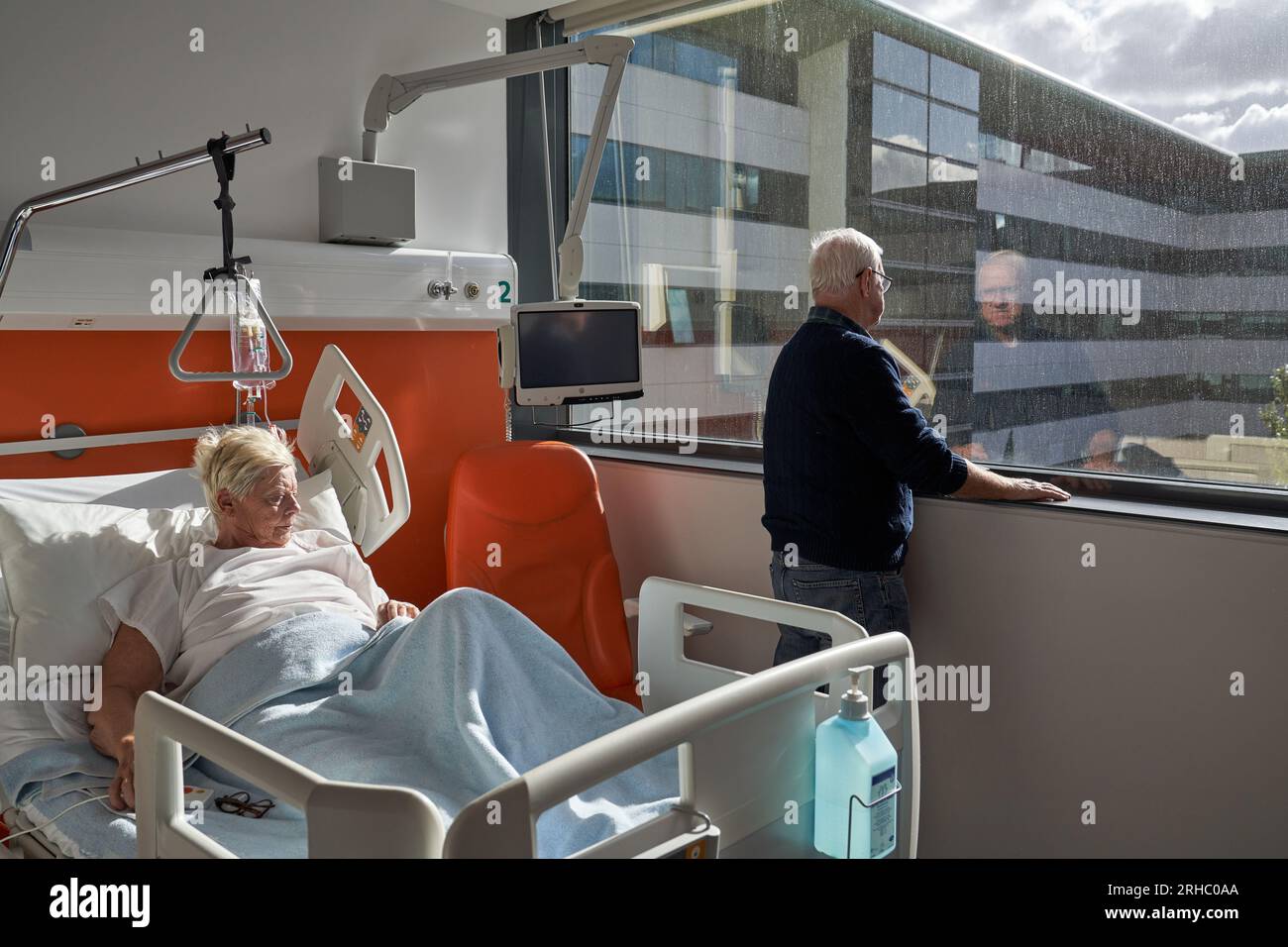 Vue latérale d'un homme âgé contrarié avec de courts cheveux blancs regardant par la fenêtre alors qu'il se tenait à l'hôpital près d'une femme malade couchée sur le canapé pendant la réhabilitation Banque D'Images