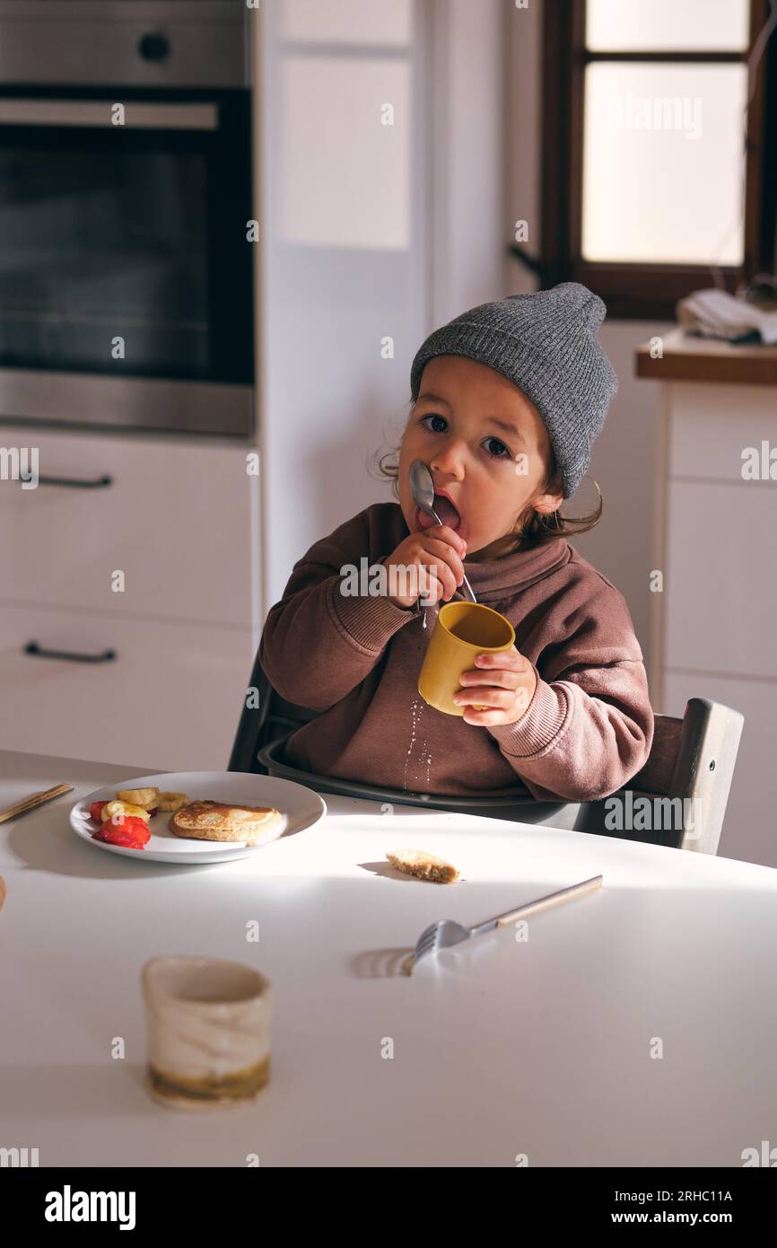 Grand angle de petit garçon souillé avec du lait renversé assis à la table et une cuillère à lécher tout en prenant le petit déjeuner avec une crêpe à la banane et aux baies Banque D'Images