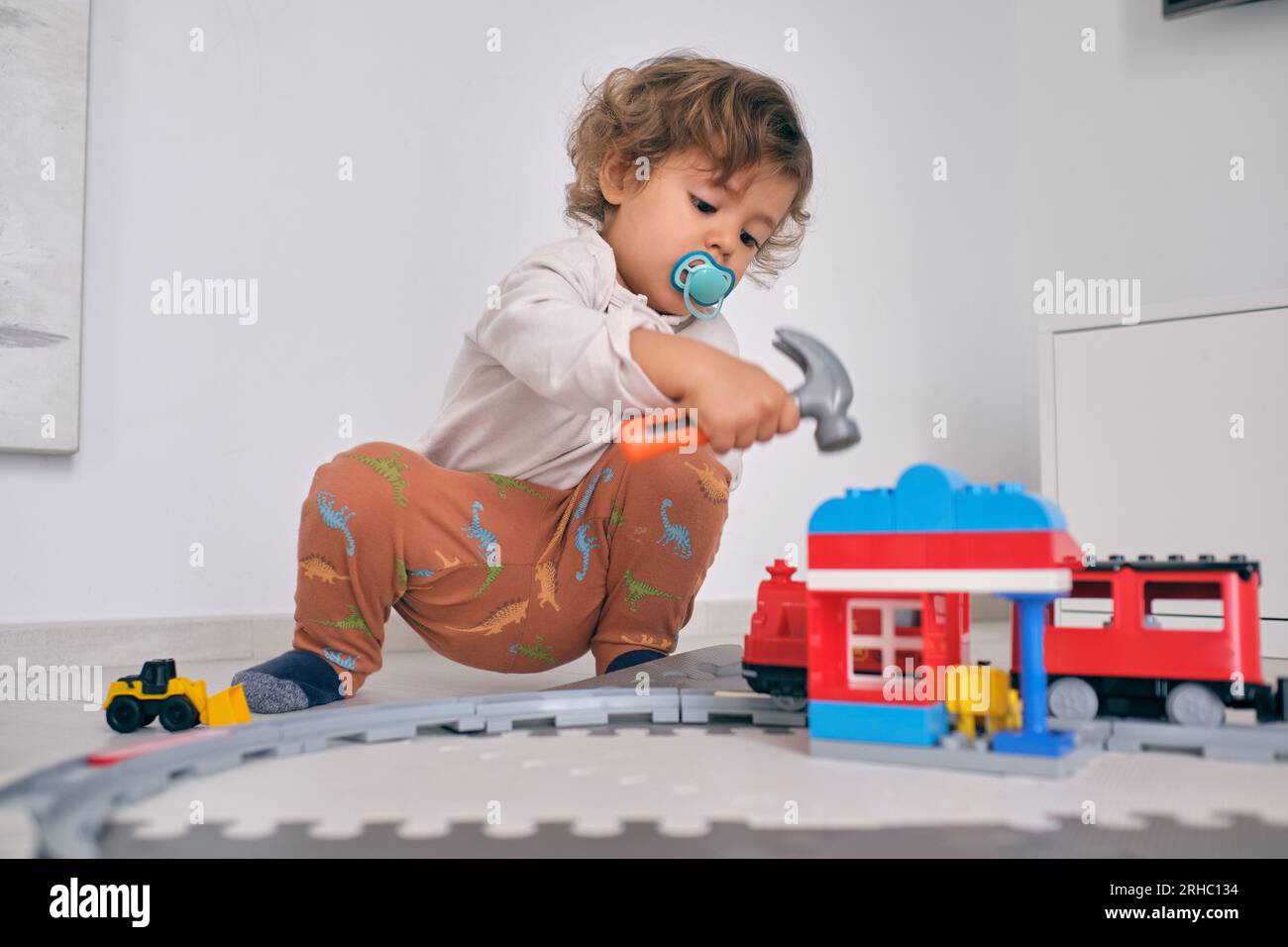 Corps complet de petit garçon curieux avec sucette assis sur le sol dans le salon et jouant avec le marteau jouet et la gare à la maison Banque D'Images