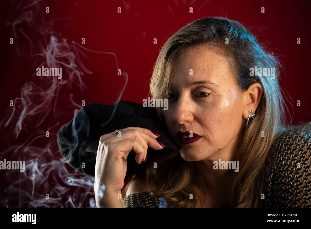 Femme attrayante et confiante tenant un béret noir à côté de son visage. Fumée d'encens tout autour. Portrait de studio sur fond rouge. Banque D'Images