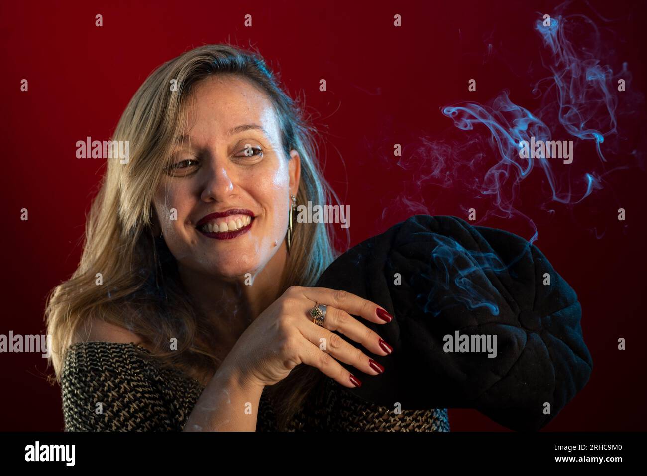 Femme attrayante et confiante tenant un béret noir à côté de son visage. Fumée d'encens tout autour. Portrait de studio sur fond rouge. Banque D'Images