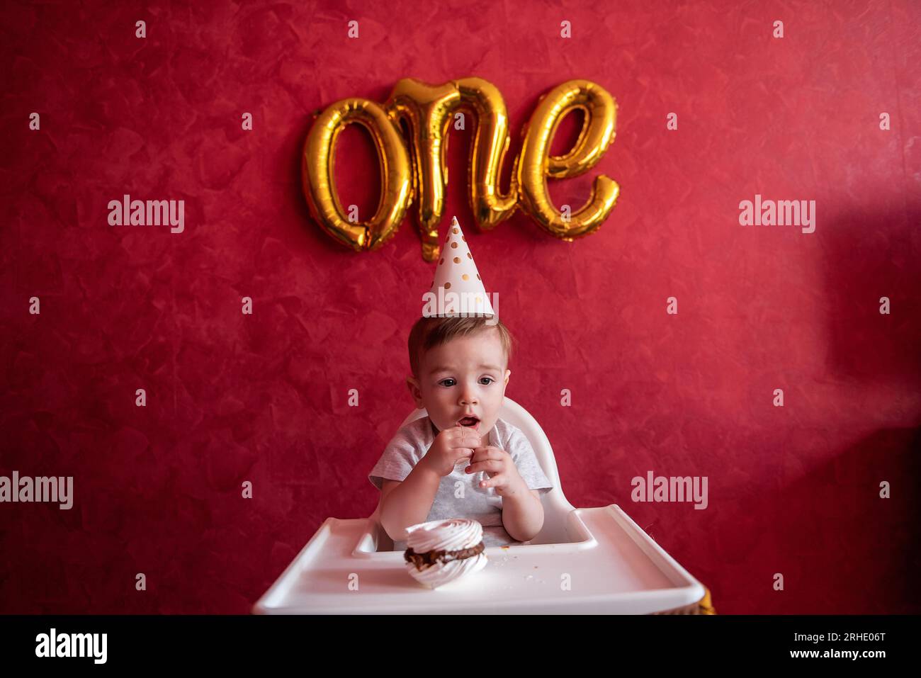 Petit garçon d'un an dans le chapeau de fête assis dans la chaise d'enfants, mangeant gâteau d'anniversaire, meringue, souffle la bougie. Kid sur fond isolé rouge, sur Banque D'Images