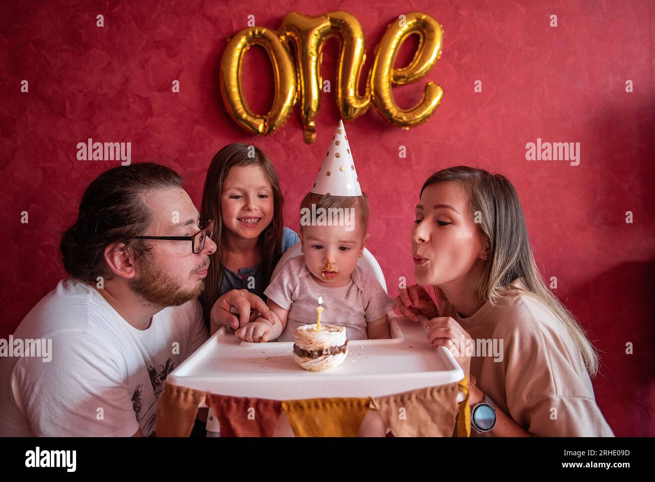 Mère et père aident à souffler la bougie à un fils d'un an sur le gâteau d'anniversaire. La sœur aînée rit. Enfant dans le chapeau de fête sur fond isolé rouge wi Banque D'Images