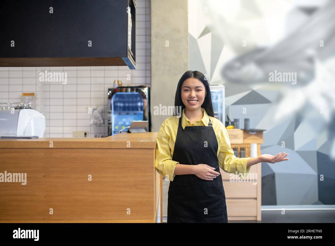 Mignonne serveuse souriante dans un tablier noir debout près du comptoir du café Banque D'Images