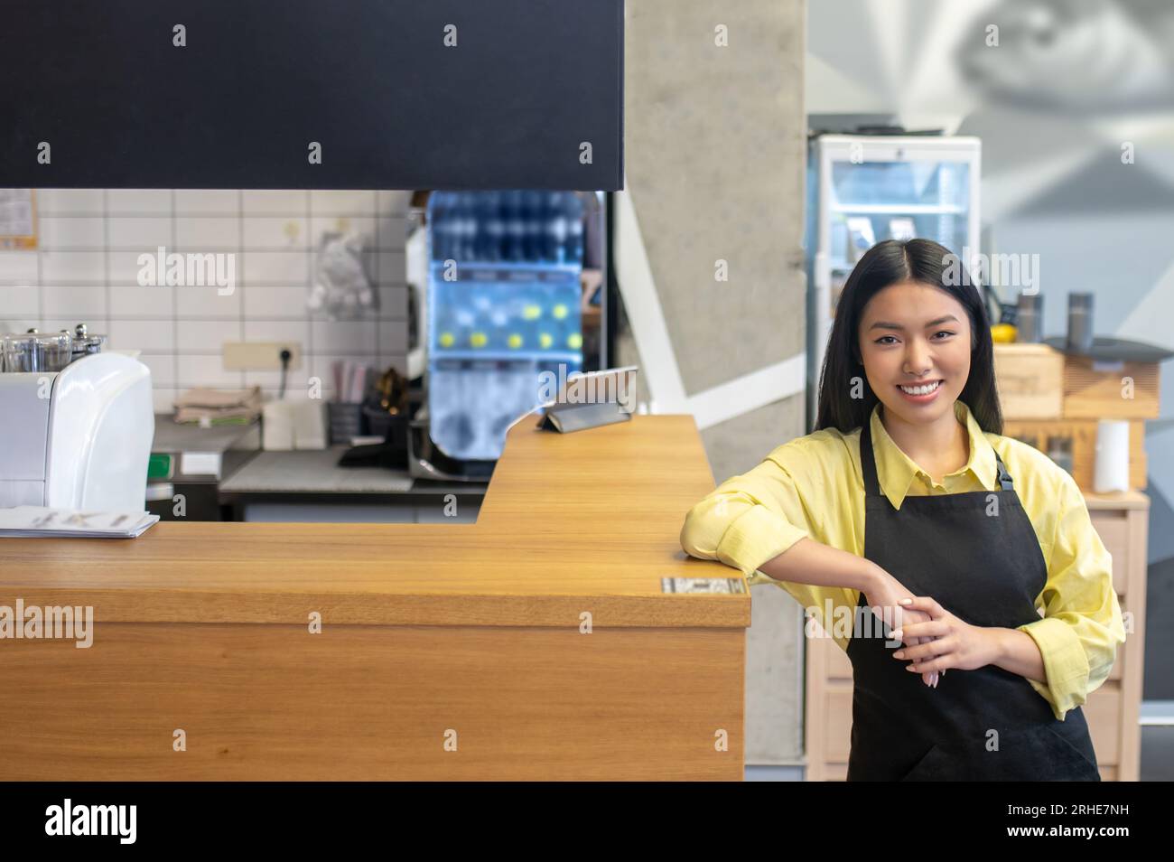 Mignonne serveuse souriante dans un tablier noir debout près du comptoir du café Banque D'Images