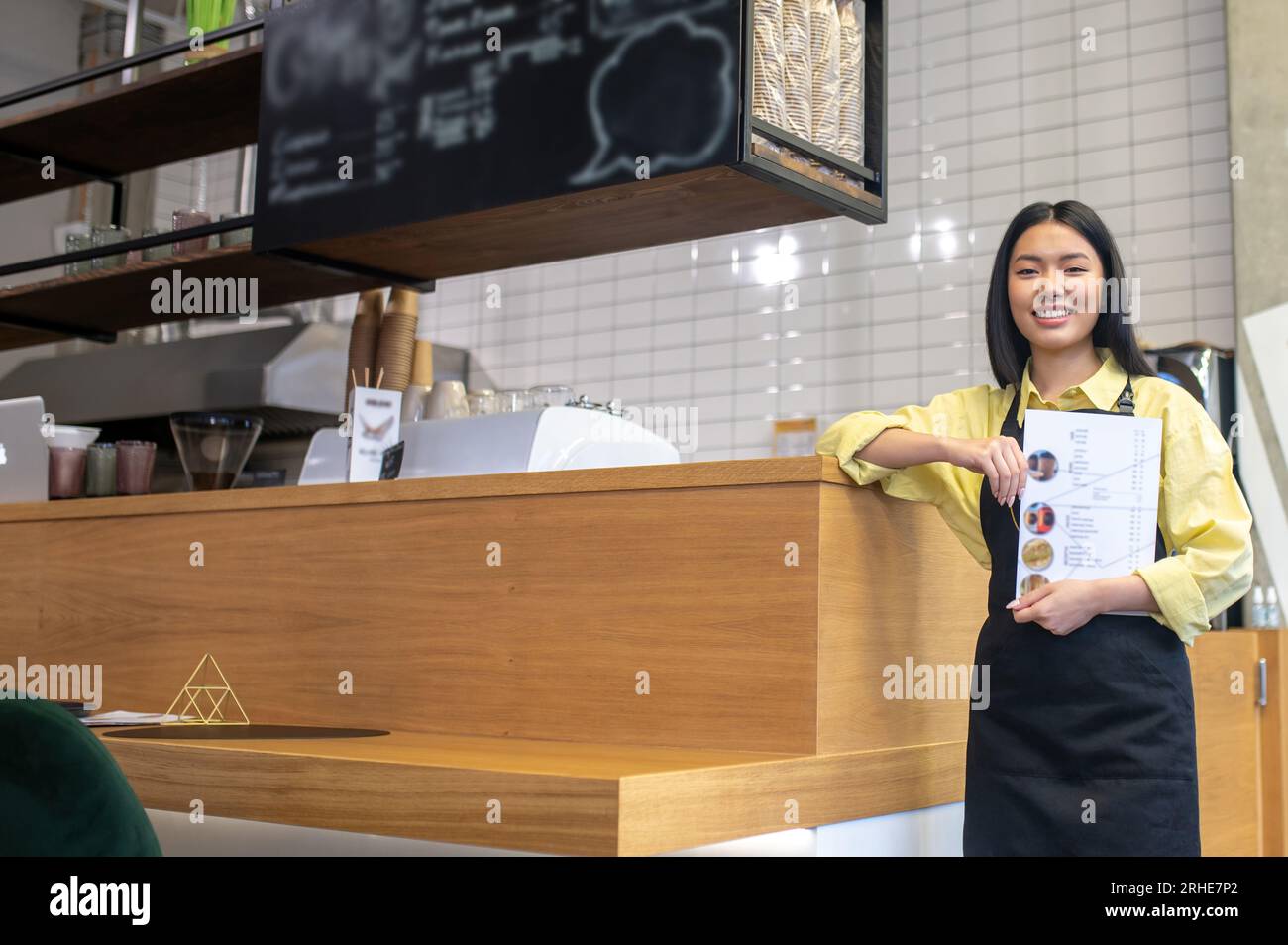 Mignonne serveuse souriante dans un tablier noir debout près du comptoir du café Banque D'Images